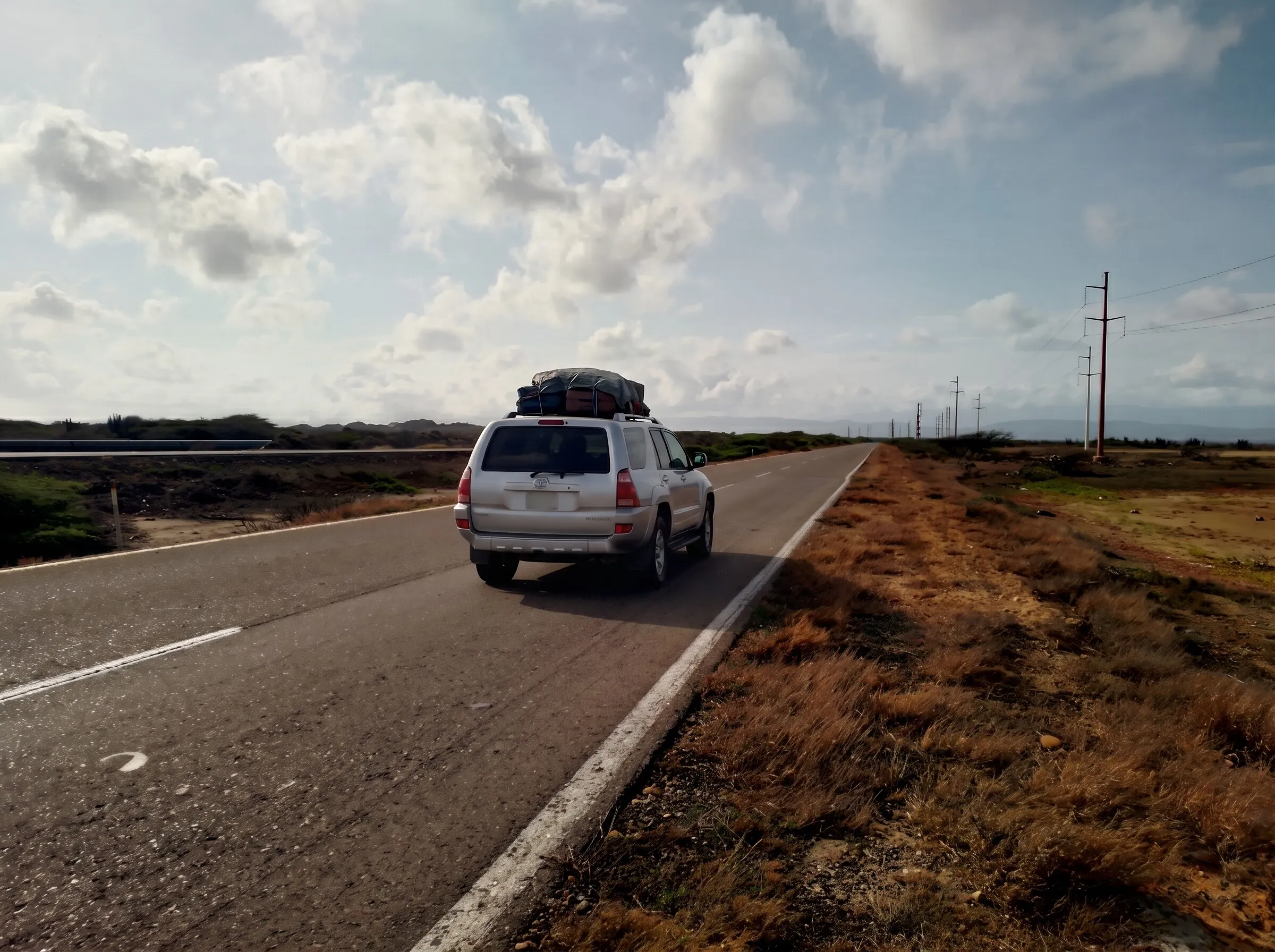 Una camioneta con maletas amarradas en el techo avanza por una carretera recta entre monte seco.