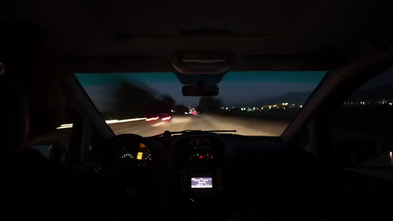 Un conductor al volante al anochecer, visto desde el asiento trasero, con la carretera iluminada al frente.