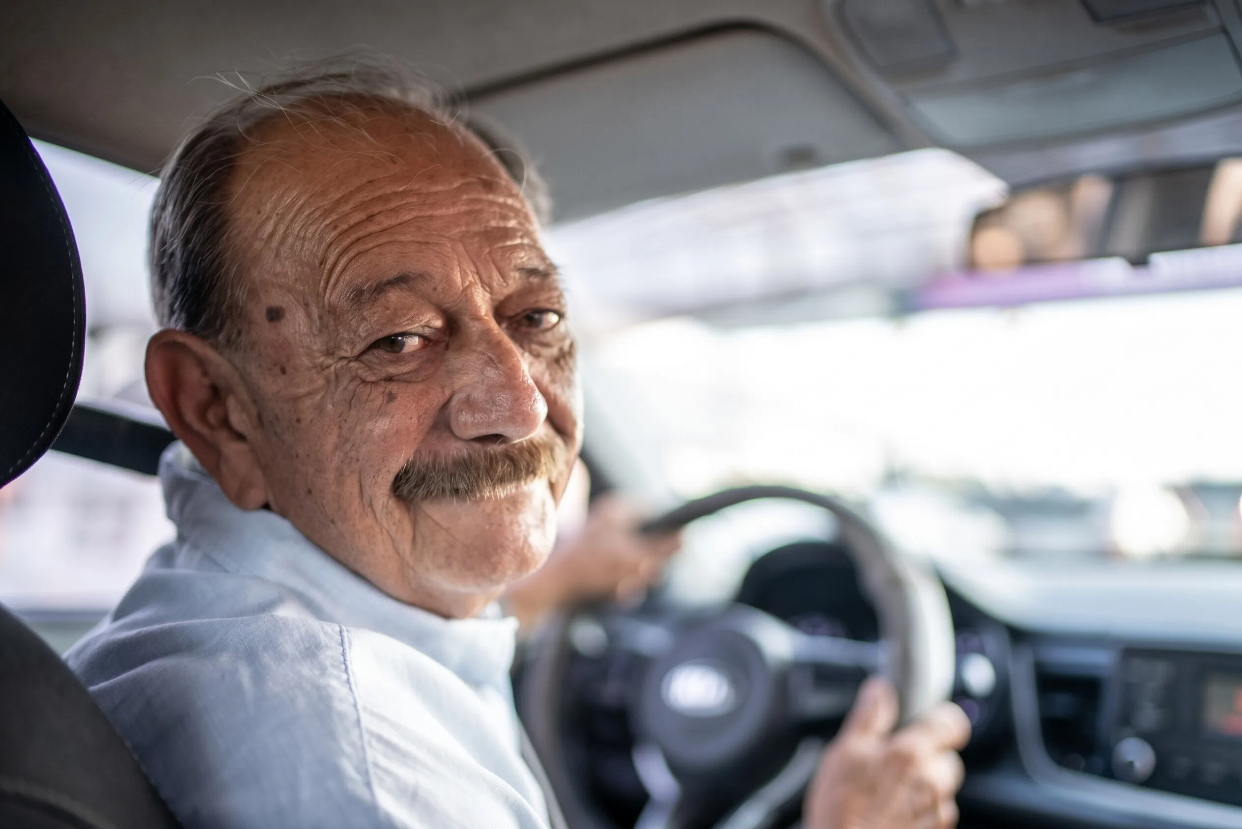 Un conductor mayor, de bigote, se voltea sonriendo hacia el pasajero con las manos en el volante.